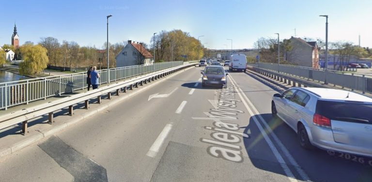 Widok z perspektywy jezdni na most Kardynała Wyszyńskiego w Elblągu. Na asfalcie widnieje namalowany napis „aleja Wyszyńskiego”. Po drodze poruszają się samochody, a po lewej stronie, na wydzielonym barierami chodniku, stoją dwie osoby. W tle po lewej widać rzekę, drzewa oraz wysoką wieżę kościoła