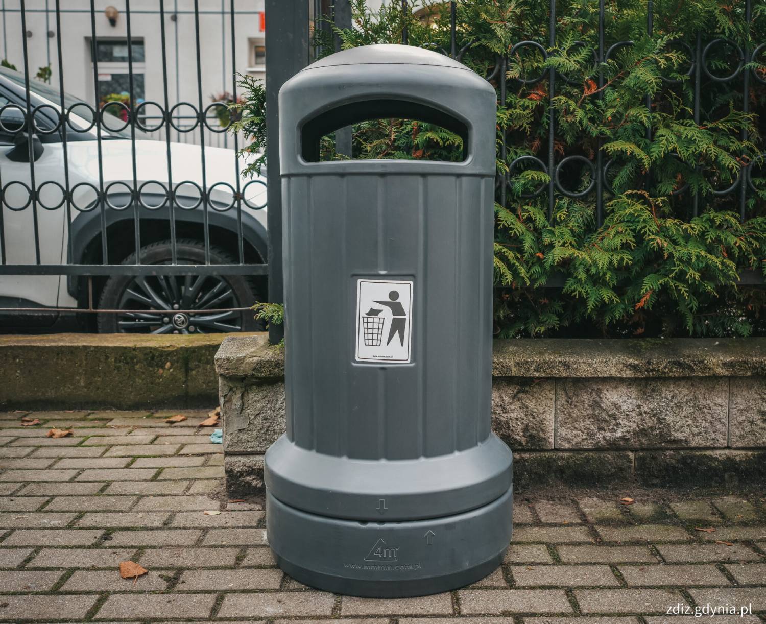 Nowoczesny, ciemnoszary, cylindryczny kosz na śmieci z zaokrągloną kopułą i szerokim otworem wrzutowym, stojący na brukowanym chodniku przy czarnym płocie i zielonych krzewach. Na środku pojemnika znajduje się biała naklejka z czarnym piktogramem przedstawiającym postać wrzucającą śmieci do kosza