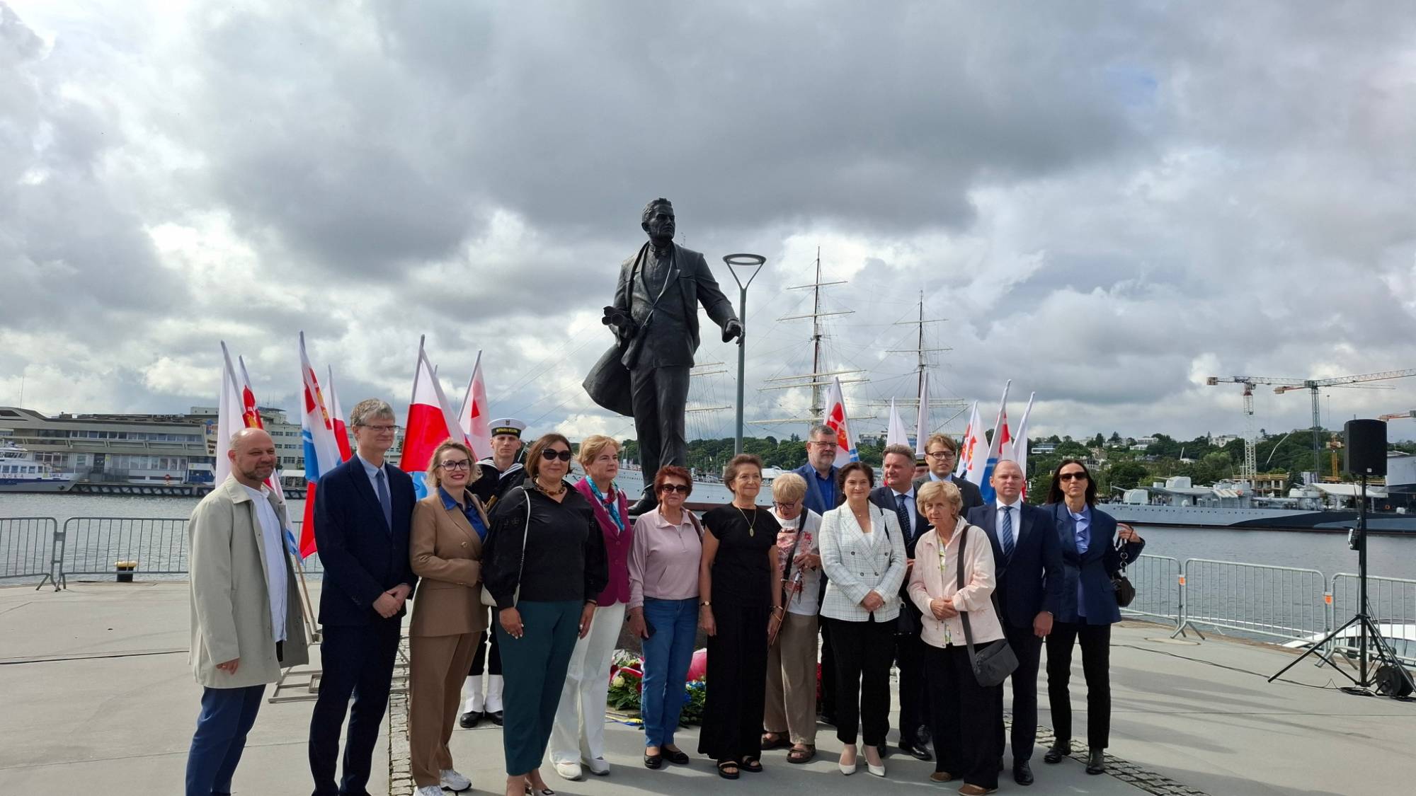 Grupowe zdjęcie uczestników uroczystości stojących w rzędzie przed pomnikiem Tadeusza Wendy w Gdyni. Grupa składająca się z kilkunastu osób - kobiet i mężczyzn w strojach wizytowych oraz jednego marynarza w tle - pozuje na tle pomnika oraz flag Polski i Gdyni. W tle widoczny jest basen portowy, maszty żaglowca, okręt wojenny oraz zachmurzone niebo