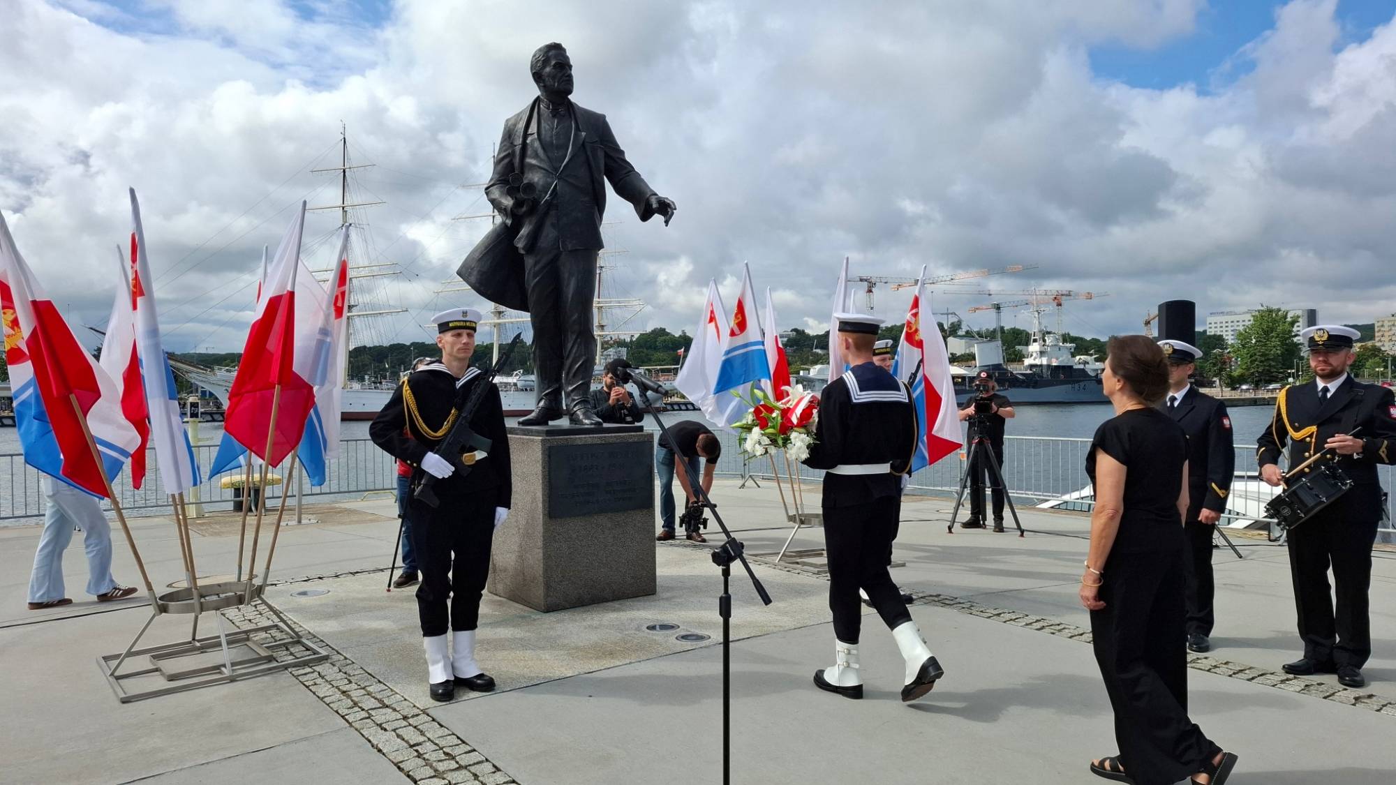 Uroczystość przed pomnikiem Tadeusza Wendy w Gdyni. Marynarz Marynarki Wojennej w galowym mundurze niesie biało-czerwony wieniec w stronę pomnika, przy którym inny marynarz stoi na warcie z bronią. Po prawej stronie wydarzenie obserwuje Hanna Wenda-Uszyńska oraz oficerowie. W tle powiewają flagi Polski i Gdyni, a w oddali widać portowe nabrzeże z jednostkami pływającymi pod zachmurzonym niebem