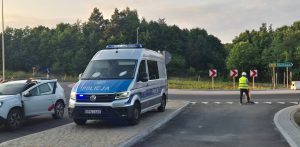 Srebrno-niebieski furgon policyjny na sygnale świetlnym, zaparkowany na nowym skrzyżowaniu obok białego auta firmy budowlanej z otwartymi drzwiami. Po prawej stronie stoi pracownik w kamizelce odblaskowej, a w tle widoczny jest drogowskaz „210 Unichowo” na tle ściany lasu. Otwarcie fragmentu słupskiego ringu
