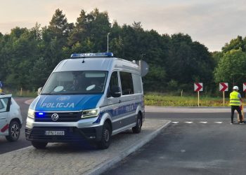 Srebrno-niebieski furgon policyjny na sygnale świetlnym, zaparkowany na nowym skrzyżowaniu obok białego auta firmy budowlanej z otwartymi drzwiami. Po prawej stronie stoi pracownik w kamizelce odblaskowej, a w tle widoczny jest drogowskaz „210 Unichowo” na tle ściany lasu. Otwarcie fragmentu słupskiego ringu