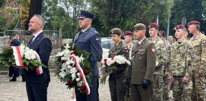 Uczestnicy obchodów 109. rocznicy uwięzienia Józefa Piłsudskiego w Gdańsku. Na pierwszym planie mężczyzna w garniturze (dr Marek Szymaniak z IPN). Uczestnicy trzymają biało-czerwone wieńce i kwiaty podczas uroczystości składania hołdu.