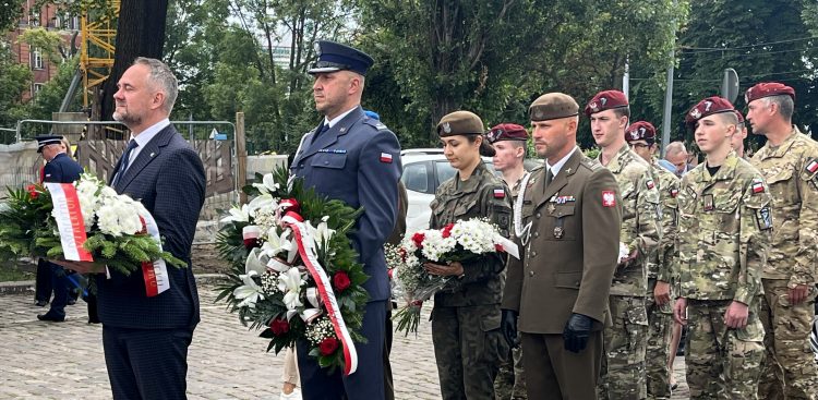 Uczestnicy obchodów 109. rocznicy uwięzienia Józefa Piłsudskiego w Gdańsku. Na pierwszym planie mężczyzna w garniturze (dr Marek Szymaniak z IPN). Uczestnicy trzymają biało-czerwone wieńce i kwiaty podczas uroczystości składania hołdu.