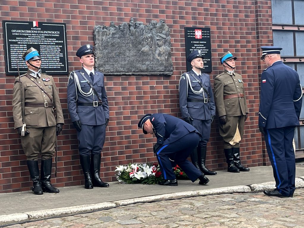 Składanie kwiatów pod tablicą upamiętniającą uwięzienie Józefa Piłsudskiego w Gdańsku podczas obchodów 109. rocznicy tego wydarzenia. Na zdjęciu widać moment oddawania hołdu przez przedstawiciela służb mundurowych w asyście warty honorowej na tle muru więziennego
