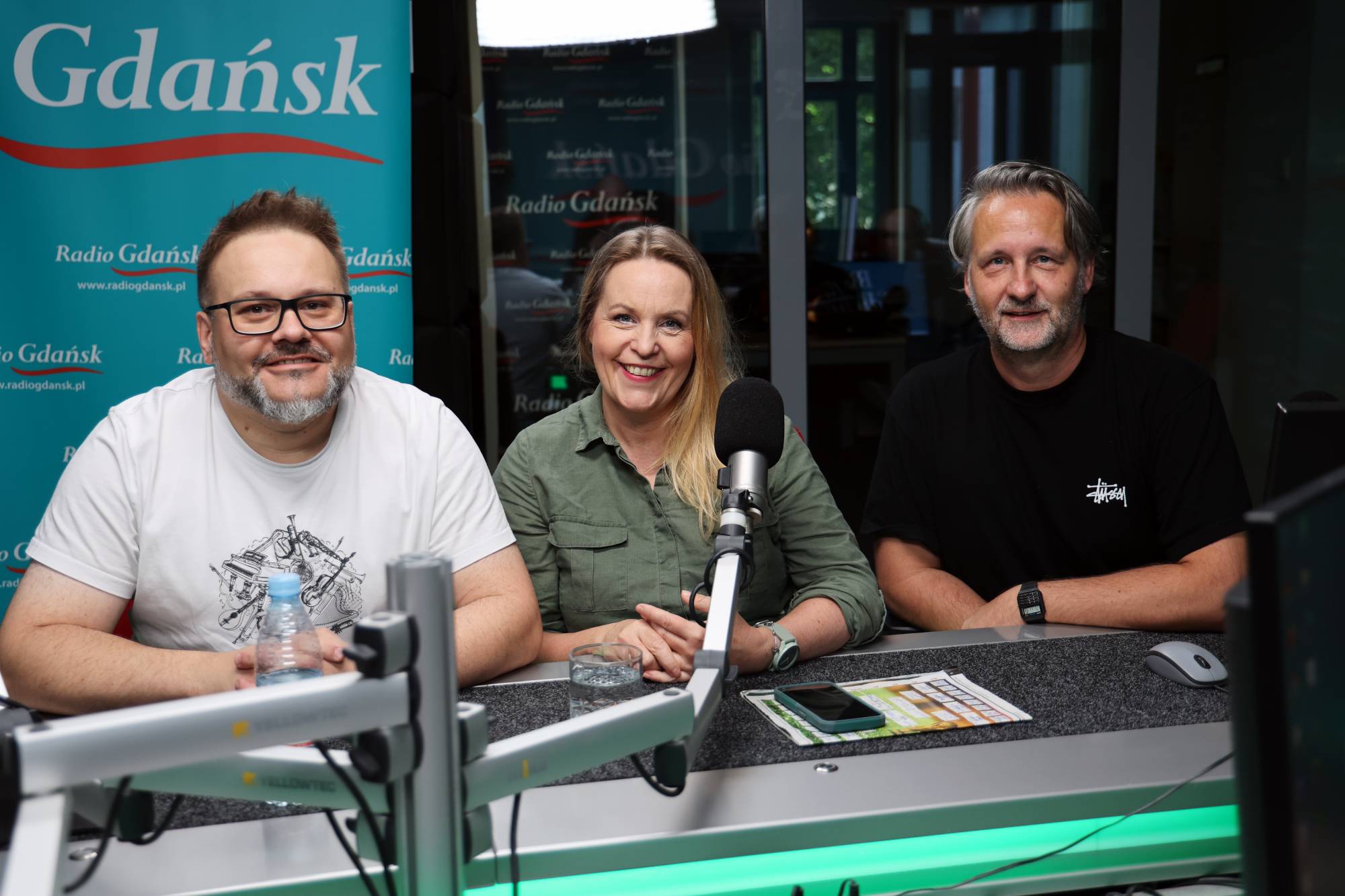 Trzy osoby – dwóch mężczyzn i kobieta – siedzą uśmiechnięci przy stole w studiu nagraniowym Radia Gdańsk. Od lewej: mężczyzna w okularach i białym t-shircie (Paweł Ryta), w środku kobieta z długimi blond włosami w oliwkowej koszuli (Katarzyna Michałowska), a po prawej mężczyzna z siwiejącym zarostem w czarnym t-shircie (Michał Stankiewicz). Przed nimi na statywach zamontowane są mikrofony studyjne, a w tle widoczna jest turkusowa ścianka z logo Radio Gdańsk