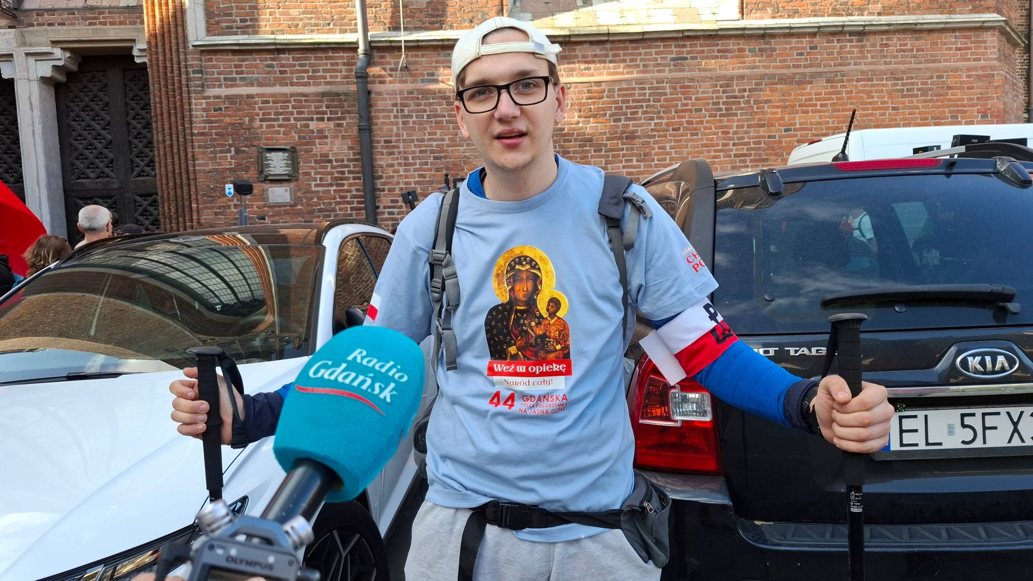 Młody mężczyzna w okularach, białej czapce z daszkiem założonej tyłem i błękitnej koszulce z wizerunkiem Matki Boskiej Częstochowskiej oraz napisem "44. Gdańska Piesza Pielgrzymka na Jasną Górę" trzyma w obu dłoniach kijki trekkingowe, a na lewym ramieniu ma biało-czerwoną Bazyliki Mariackiej w Gdańsku