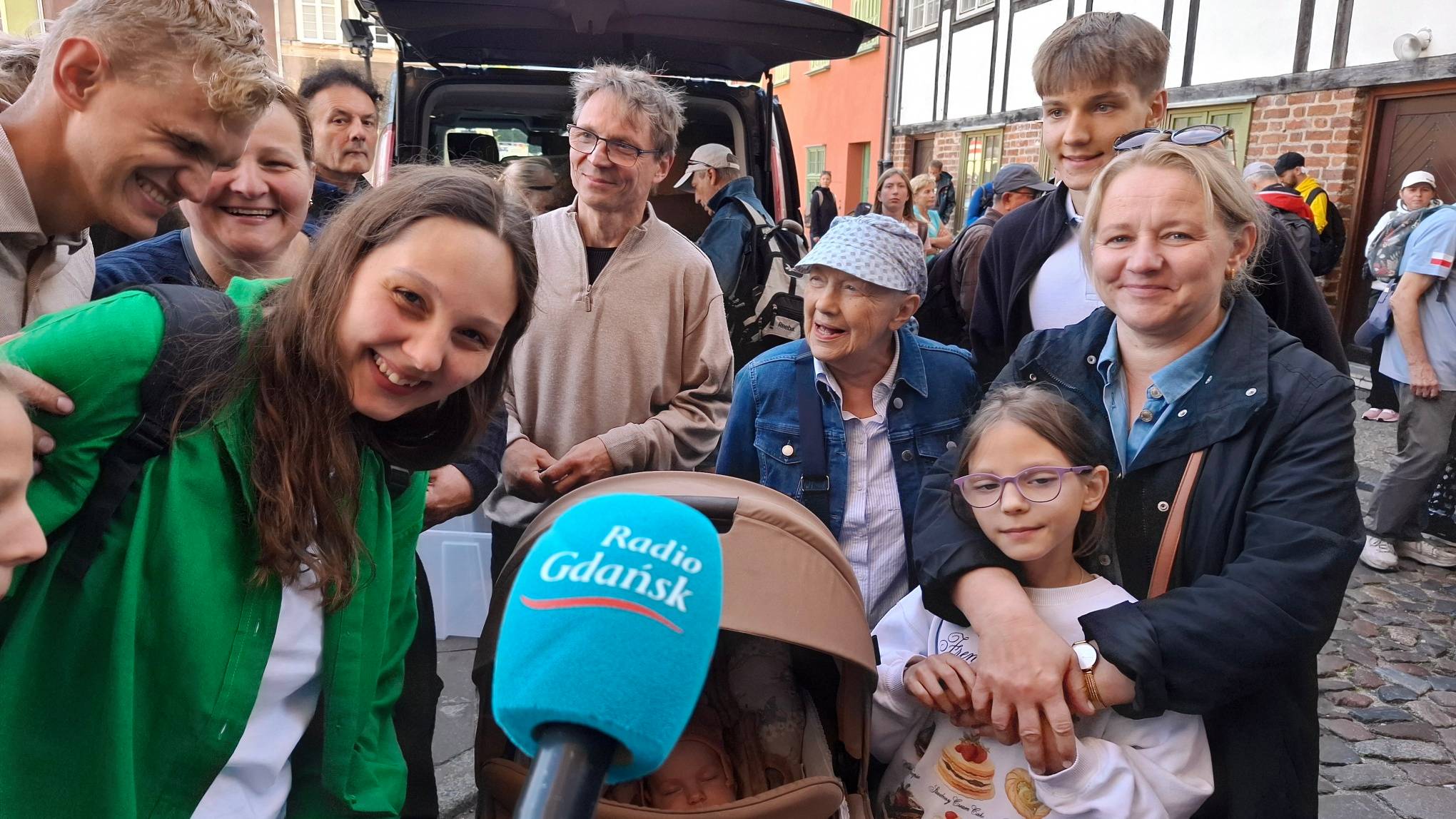 Pielgrzymi ruszają z Gdańska na Jasną Górę. Uśmiechnięta, wielopokoleniowa rodzina pozuje wokół beżowego wózka dziecięcego, w którym śpi niemowlę. Na pierwszym planie widać niebieski mikrofon z logo "Radio Gdańsk". Po prawej stronie kobieta obejmuje kilkuletnią dziewczynkę w okularach, obok nich stoi starsza kobieta w czapce i dżinsowej kurtce, a po lewej młodsi i starsi członkowie rodziny pochylają się z uśmiechem w stronę wózka. W tle widoczny jest otwarty bagażnik samochodu oraz zabytkowa zabudowa