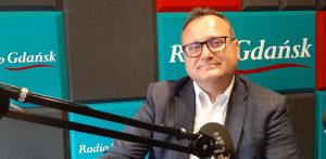 Mężczyzna w okularach, białej koszuli i ciemnej marynarce siedzi w studiu nagraniowym, patrząc w stronę obiektywu. Przed nim widoczny jest profesjonalny mikrofon radiowy na statywie. W tle znajdują się ścienne panele akustyczne w kolorze turkusowym i czerwonym z widocznym białym logotypem „Radio Gdańsk”. To Piotr Rachwalski, członek zarządu spółki CPK Port Polska