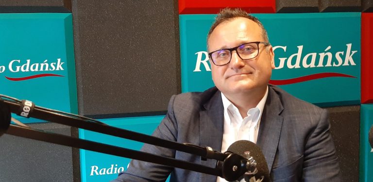 Mężczyzna w okularach, białej koszuli i ciemnej marynarce siedzi w studiu nagraniowym, patrząc w stronę obiektywu. Przed nim widoczny jest profesjonalny mikrofon radiowy na statywie. W tle znajdują się ścienne panele akustyczne w kolorze turkusowym i czerwonym z widocznym białym logotypem „Radio Gdańsk”. To Piotr Rachwalski, członek zarządu spółki CPK Port Polska