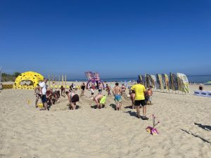 Plażowe Mistrzostwa Budowniczych. Szerokie ujęcie słonecznej plaży, na której grupa ludzi w strojach kąpielowych i letnich ubraniach wspólnie buduje górę z piasku. W prawym dolnym rogu na piasku leży różowa zabawkowa łopatka. W tle, po lewej stronie, stoi duży żółty namiot sferyczny w czarne plamy przypominający ser, pośrodku fioletowy łuk reklamowy, a po prawej rząd pionowych flag plażowych. W oddali widać morze pod bezchmurnym, błękitnym niebem
