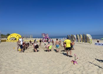 Plażowe Mistrzostwa Budowniczych. Szerokie ujęcie słonecznej plaży, na której grupa ludzi w strojach kąpielowych i letnich ubraniach wspólnie buduje górę z piasku. W prawym dolnym rogu na piasku leży różowa zabawkowa łopatka. W tle, po lewej stronie, stoi duży żółty namiot sferyczny w czarne plamy przypominający ser, pośrodku fioletowy łuk reklamowy, a po prawej rząd pionowych flag plażowych. W oddali widać morze pod bezchmurnym, błękitnym niebem