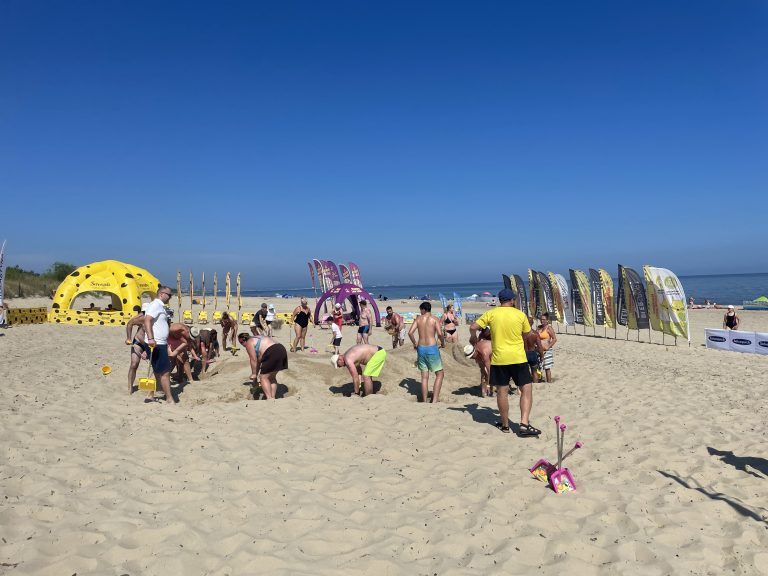 Plażowe Mistrzostwa Budowniczych. Szerokie ujęcie słonecznej plaży, na której grupa ludzi w strojach kąpielowych i letnich ubraniach wspólnie buduje górę z piasku. W prawym dolnym rogu na piasku leży różowa zabawkowa łopatka. W tle, po lewej stronie, stoi duży żółty namiot sferyczny w czarne plamy przypominający ser, pośrodku fioletowy łuk reklamowy, a po prawej rząd pionowych flag plażowych. W oddali widać morze pod bezchmurnym, błękitnym niebem