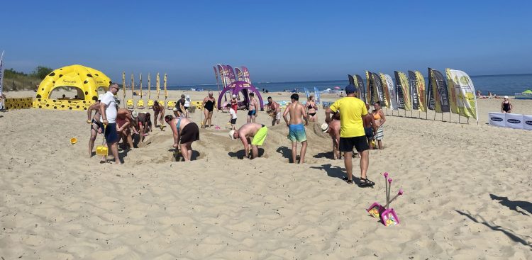 Plażowe Mistrzostwa Budowniczych. Szerokie ujęcie słonecznej plaży, na której grupa ludzi w strojach kąpielowych i letnich ubraniach wspólnie buduje górę z piasku. W prawym dolnym rogu na piasku leży różowa zabawkowa łopatka. W tle, po lewej stronie, stoi duży żółty namiot sferyczny w czarne plamy przypominający ser, pośrodku fioletowy łuk reklamowy, a po prawej rząd pionowych flag plażowych. W oddali widać morze pod bezchmurnym, błękitnym niebem
