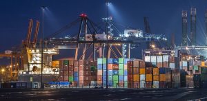Oświetlony nocą terminal kontenerowy w porcie z rzędami wielobarwnych kontenerów, wysokimi suwnicami bramowymi z logo GCT oraz fragmentem zacumowanego statku handlowego „Thetis D”