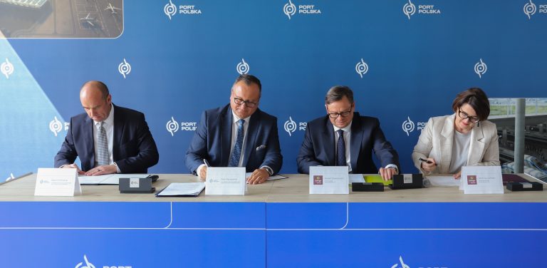 Cztery osoby – trzej mężczyźni i kobieta w jasnym żakiecie – siedzą obok siebie przy długim stole i podpisują dokumenty. W tle znajduje się niebieska ścianka z powtarzającym się białym logo „PORT POLSKA”. Przed każdą z osób na stole ustawiona jest biała tabliczka informacyjna. Moment podpisania porozumienia pomiędzy przedstawicielami Port Polska i Energa-Operator dotyczącego koordynacji inwestycji kolejowych i energetycznych