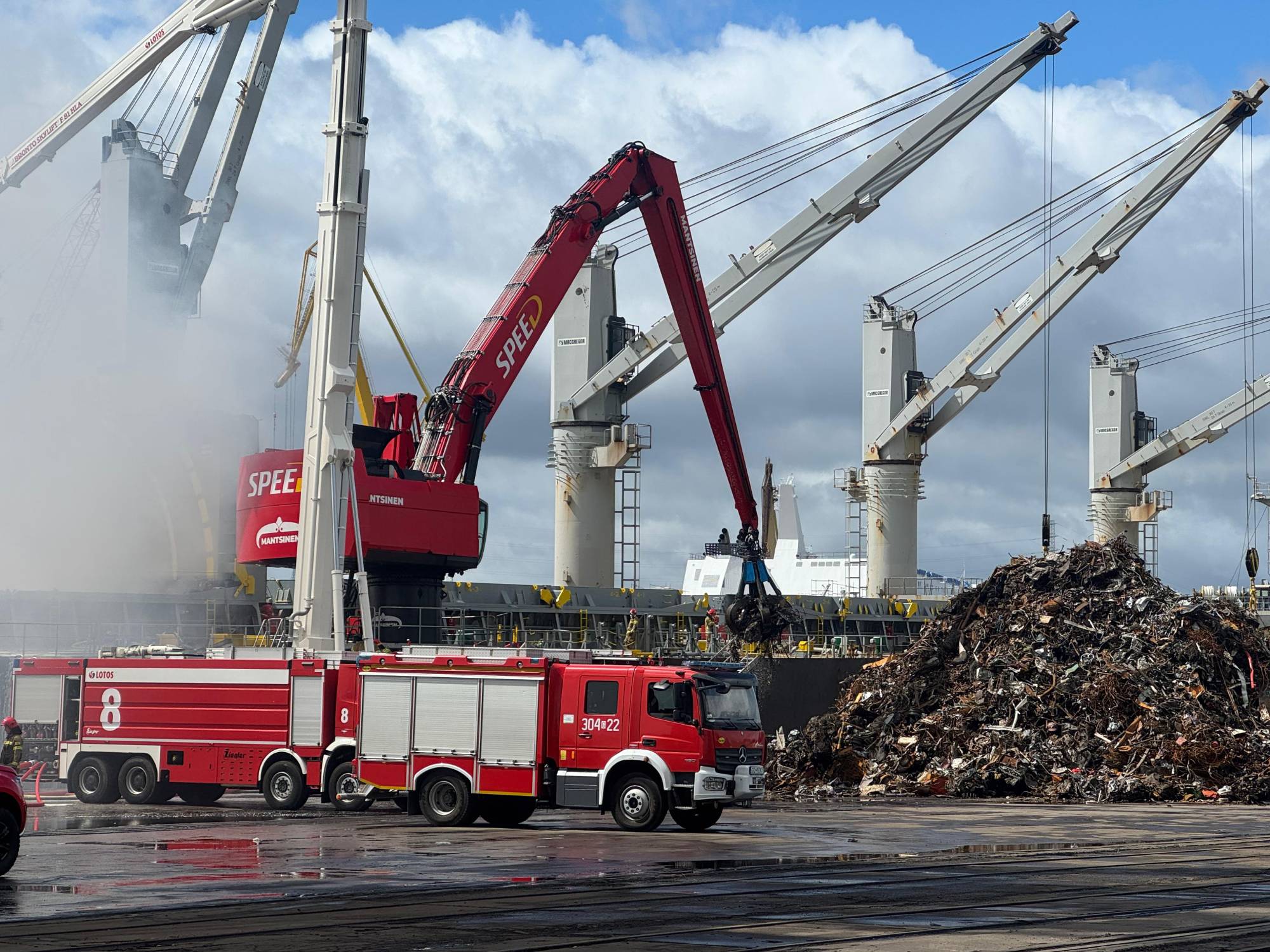 Pożar statku w Porcie Gdańsk. Dźwig przeładowuje złom ze statku na nabrzeże. Widać dym i wozy strażackie, na drugim planie szare portowe dźwigi przeładunkowe