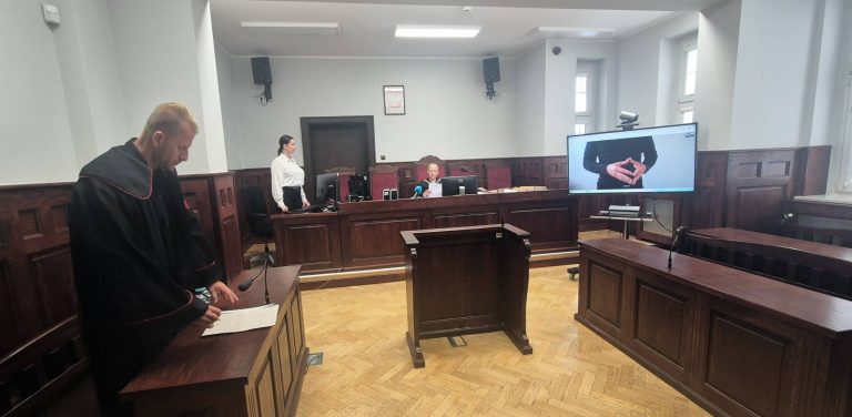 Szerokie ujęcie sali rozpraw Sądu Okręgowego w Słupsku. Po lewej stronie stoi prokurator w todze z czerwonymi elementami, przeglądający dokumenty. W głębi, za drewnianym stołem sędziowskim, siedzi sędzia w todze, a obok stoi protokolantka. Po prawej stronie na dużym ekranie telewizora wyświetlane jest połączenie wideo z oskarżonym przebywającym w zakładzie karnym. Sąd skazał Piotra S. na karę trzech lat więzienia za oszustwo - miał wyłudzić pieniądze na instalację do przetwarzania odpadów gumowych, z czego się nie wywiązał