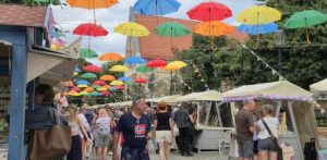 Jarmark św. Dominika w Gdańsku. Brukowany deptak jarmarczny, nad którym na linach zawieszono dziesiątki otwartych, kolorowych parasoli w barwach czerwonej, pomarańczowej, żółtej, zielonej i niebieskiej. Po obu stronach uliczki stoją drewniane kramy z jasnymi zadaszeniami, a między nimi spacerują ludzie w letnich ubraniach. W tle widać drzewa oraz fragment budynku z czerwonym dachem