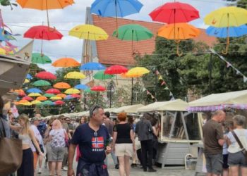 Jarmark św. Dominika w Gdańsku. Brukowany deptak jarmarczny, nad którym na linach zawieszono dziesiątki otwartych, kolorowych parasoli w barwach czerwonej, pomarańczowej, żółtej, zielonej i niebieskiej. Po obu stronach uliczki stoją drewniane kramy z jasnymi zadaszeniami, a między nimi spacerują ludzie w letnich ubraniach. W tle widać drzewa oraz fragment budynku z czerwonym dachem