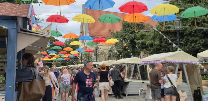 Jarmark św. Dominika w Gdańsku. Brukowany deptak jarmarczny, nad którym na linach zawieszono dziesiątki otwartych, kolorowych parasoli w barwach czerwonej, pomarańczowej, żółtej, zielonej i niebieskiej. Po obu stronach uliczki stoją drewniane kramy z jasnymi zadaszeniami, a między nimi spacerują ludzie w letnich ubraniach. W tle widać drzewa oraz fragment budynku z czerwonym dachem