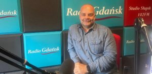 Uśmiechnięty, łysy mężczyzna w niebieskiej koszuli jeansowej siedzi w studiu radiowym. W tle widoczne są turkusowe i czerwone panele akustyczne z napisami „Radio Gdańsk” oraz „Studio Słupsk 102FM”. Na pierwszym planie, z boku, znajduje się profesjonalny mikrofon studyjny. To psycholog Maciej Maraszkiewicz
