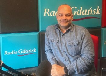 Uśmiechnięty, łysy mężczyzna w niebieskiej koszuli jeansowej siedzi w studiu radiowym. W tle widoczne są turkusowe i czerwone panele akustyczne z napisami „Radio Gdańsk” oraz „Studio Słupsk 102FM”. Na pierwszym planie, z boku, znajduje się profesjonalny mikrofon studyjny. To psycholog Maciej Maraszkiewicz