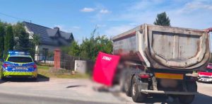 Na drodze obok domu jednorodzinnego stoi duża ciężarówka (wywrotka). Z lewej strony pojazdu rozstawiony jest czerwony parawan ochronny z białym napisem „STRAŻ”. Po lewej stronie zdjęcia, na poboczu, zaparkowany jest radiowóz policyjny z włączonymi sygnałami świetlnymi. Część kadru wokół parawanu jest celowo rozmyta