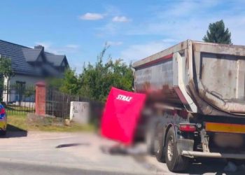 Na drodze obok domu jednorodzinnego stoi duża ciężarówka (wywrotka). Z lewej strony pojazdu rozstawiony jest czerwony parawan ochronny z białym napisem „STRAŻ”. Po lewej stronie zdjęcia, na poboczu, zaparkowany jest radiowóz policyjny z włączonymi sygnałami świetlnymi. Część kadru wokół parawanu jest celowo rozmyta