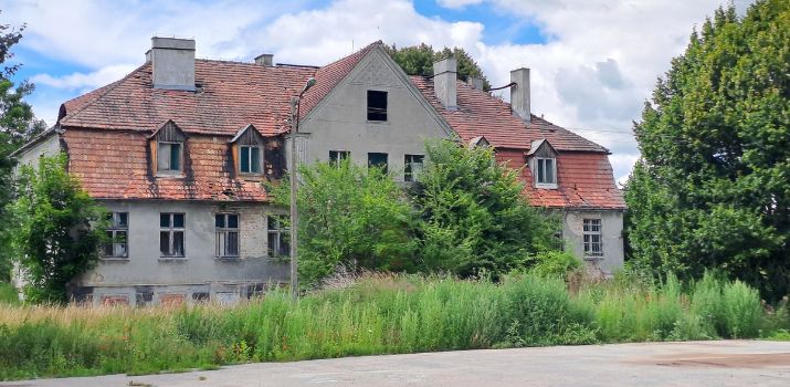 Zaniedbany, dwupiętrowy pałac w Boboszewie z niszczejącą szarą elewacją i uszkodzonym dachem z czerwonej dachówki, otoczony gęstymi, zielonymi zaroślami i wysoką trawą pod zachmurzonym niebem. W budynku mają powstać mieszkania treningowe i wspomagane