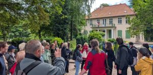 Spacer z przewodnikiem w ramach projektu "Zakamarki Gdańskich Przedmieść". Grupa turystów i mieszkańców uczestniczących w spacerze z przewodnikiem stoi na brukowanym placu przed zabytkowym Dworem Oruńskim w Gdańsku. Klasycystyczny, beżowy budynek o czerwonym, czterospadowym dachu posiada zielone okiennice na parterze oraz portyk wsparty na białych kolumnach, nad którym znajduje się balkon z polską flagą. Zgromadzeni ludzie, ubrani w codzienne stroje turystyczne, patrzą w stronę dworku, a jeden z mężczyzn na pierwszym planie unosi telefon, by zrobić zdjęcie