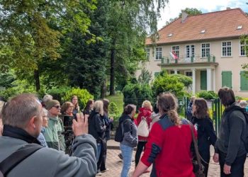 Spacer z przewodnikiem w ramach projektu "Zakamarki Gdańskich Przedmieść". Grupa turystów i mieszkańców uczestniczących w spacerze z przewodnikiem stoi na brukowanym placu przed zabytkowym Dworem Oruńskim w Gdańsku. Klasycystyczny, beżowy budynek o czerwonym, czterospadowym dachu posiada zielone okiennice na parterze oraz portyk wsparty na białych kolumnach, nad którym znajduje się balkon z polską flagą. Zgromadzeni ludzie, ubrani w codzienne stroje turystyczne, patrzą w stronę dworku, a jeden z mężczyzn na pierwszym planie unosi telefon, by zrobić zdjęcie