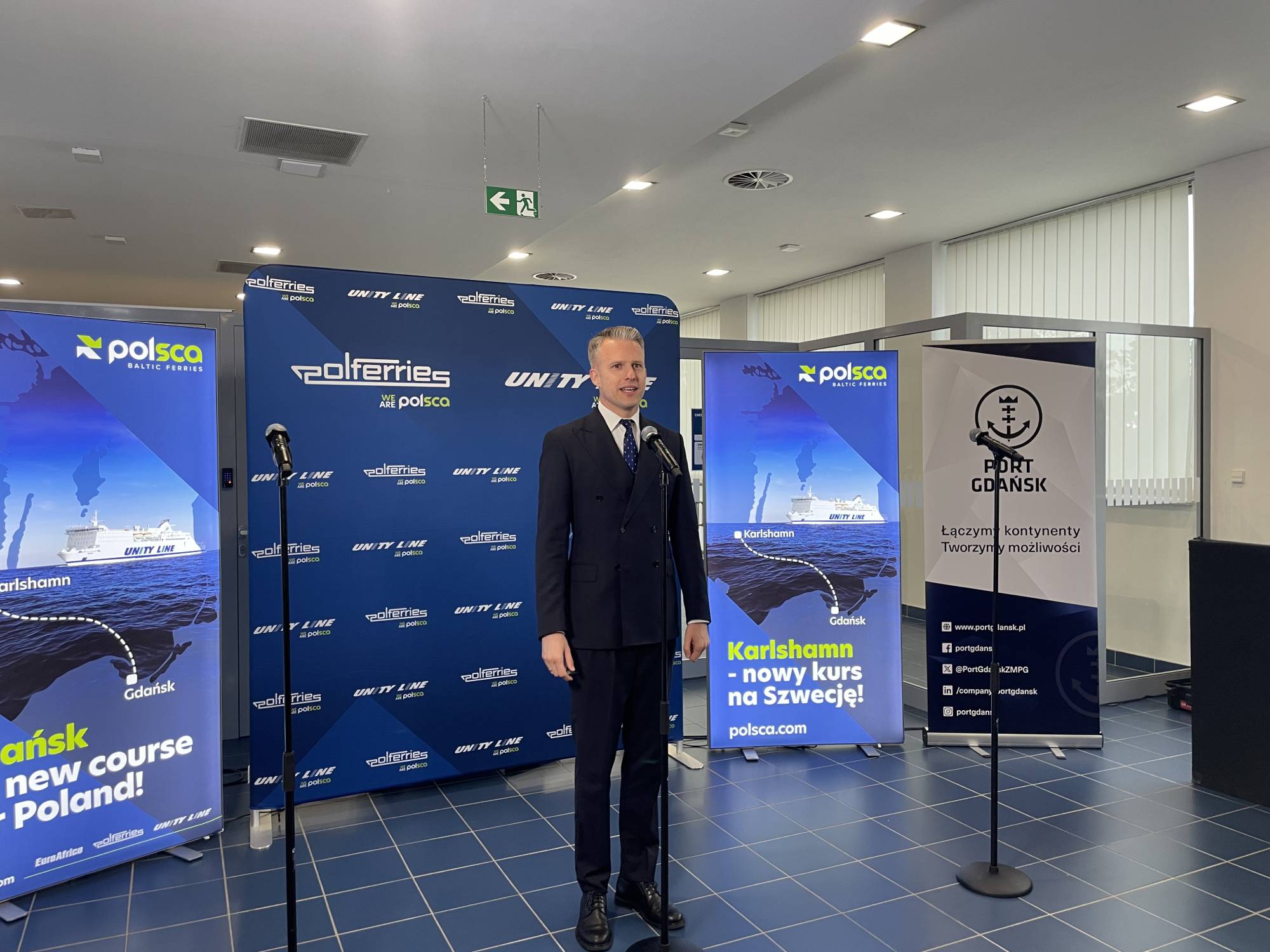 Inauguracja nowego połączenia promowego Gdańsk - Karlshamn (Szwecja). Wiceminister infrastruktury Arkadiusz Marchewka w ciemnym, dwurzędowym garniturze przemawia do mikrofonu na stojaku. Stoi w jasnym pomieszczeniu o niebieskich kafelkach na podłodze, na tle ścianki reklamowej z logo marek Polferries, Unity Line i Polsca. Po bokach widoczne są banery informujące o nowym połączeniu „Karlshamn – nowy kurs na Szwecję!” oraz oficjalny baner Portu Gdańsk