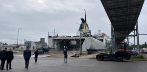 Nowe połączenie promowe Gdańsk - Karlshamn (Szwecja). Biało-niebieski prom pasażersko-samochodowy z żółto-niebieskim kominem, zacumowany przy terminalu w Porcie Gdańsk z otwartą rampą rufową. Po lewej stronie kadru idzie kilku mężczyzn w ciemnych garniturach, pośrodku stoi tyłem mężczyzna fotografujący prom smartfonem, a po prawej widoczny jest czerwony ciągnik terminalowy stojący pod metalową kładką dla pieszych. Niebo nad portem jest całkowicie zachmurzone.