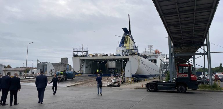 Nowe połączenie promowe Gdańsk - Karlshamn (Szwecja). Biało-niebieski prom pasażersko-samochodowy z żółto-niebieskim kominem, zacumowany przy terminalu w Porcie Gdańsk z otwartą rampą rufową. Po lewej stronie kadru idzie kilku mężczyzn w ciemnych garniturach, pośrodku stoi tyłem mężczyzna fotografujący prom smartfonem, a po prawej widoczny jest czerwony ciągnik terminalowy stojący pod metalową kładką dla pieszych. Niebo nad portem jest całkowicie zachmurzone.