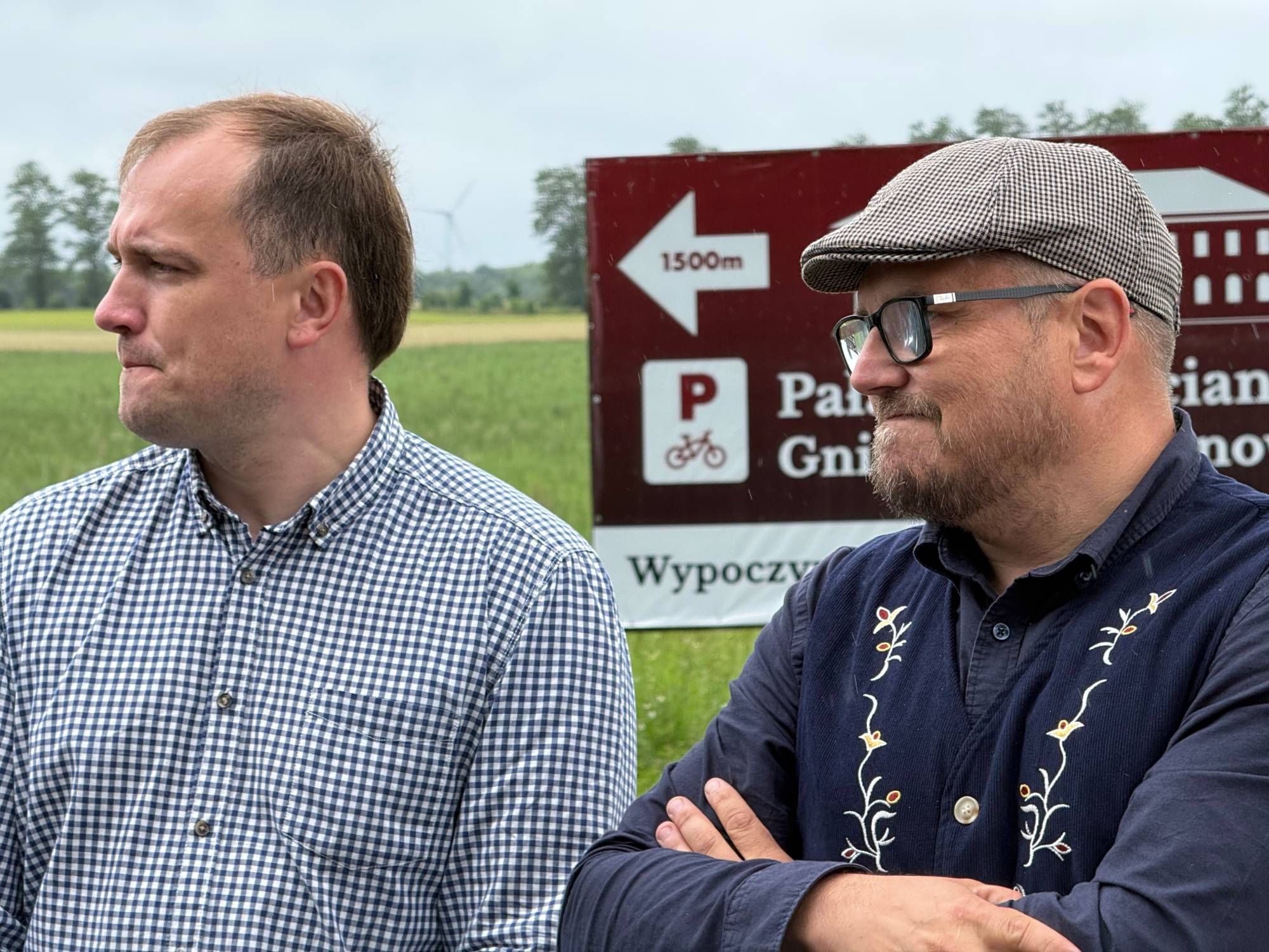 Bliskie ujęcie dwóch mężczyzn spoglądających w lewą stronę z zatroskanymi minami. Mężczyzna po lewej ubrany jest w niebiesko-białą koszulę w kratkę. Mężczyzna po prawej nosi kaszkiet, okulary i granatową kamizelkę z tradycyjnym haftem roślinnym. W tle za nimi widoczna jest brązowa tablica informacyjna z symbolem parkingu oraz napisem wskazującym dojazd do Pałacu pod Bocianim Gniazdem w Runowie. To przedsiębiorcy mający biznesy przy starej DK6 między Słupskiem a Lęborkiem, którzy tracą możliwe zyski przez błąd GPS utrudniający dojazd turystom 