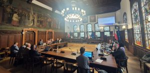 Szerokie ujęcie zabytkowej sali obrad Rady Miejskiej w Słupsku podczas sesji. Radni siedzą przy połączonych w podkowę drewnianych stołach konferencyjnych z laptopami i mikrofonami. Po lewej stronie widoczne jest monumentalne, historyczne malowidło ścienne, po prawej wysokie, zdobione witraże, a u sufitu wisi okazały, wieloramienny żyrandol. W głębi sali znajduje się stół prezydialny oraz ekran projekcyjny