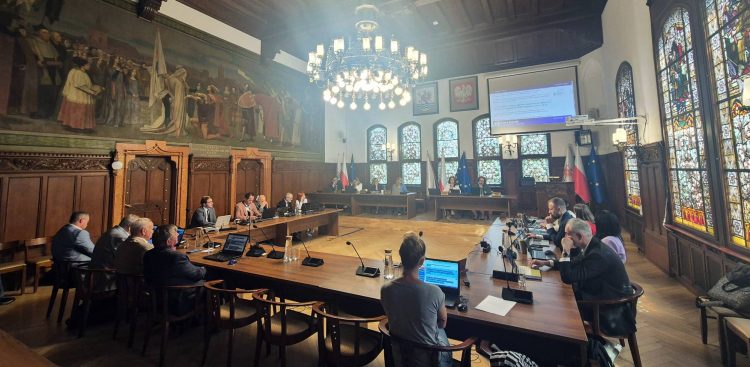 Szerokie ujęcie zabytkowej sali obrad Rady Miejskiej w Słupsku podczas sesji. Radni siedzą przy połączonych w podkowę drewnianych stołach konferencyjnych z laptopami i mikrofonami. Po lewej stronie widoczne jest monumentalne, historyczne malowidło ścienne, po prawej wysokie, zdobione witraże, a u sufitu wisi okazały, wieloramienny żyrandol. W głębi sali znajduje się stół prezydialny oraz ekran projekcyjny