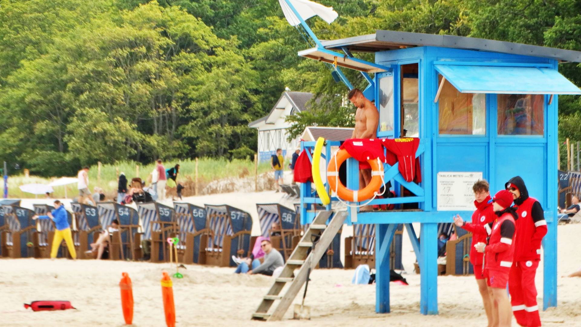Niebieska wieża ratownicza na plaży w Ustce z powiewającą białą flagą. Na podeście stoi ratownik, a na dole czuwa trzech ratowników w czerwonych kurtkach. W tle widać pasiaste kosze plażowe oraz zielony las