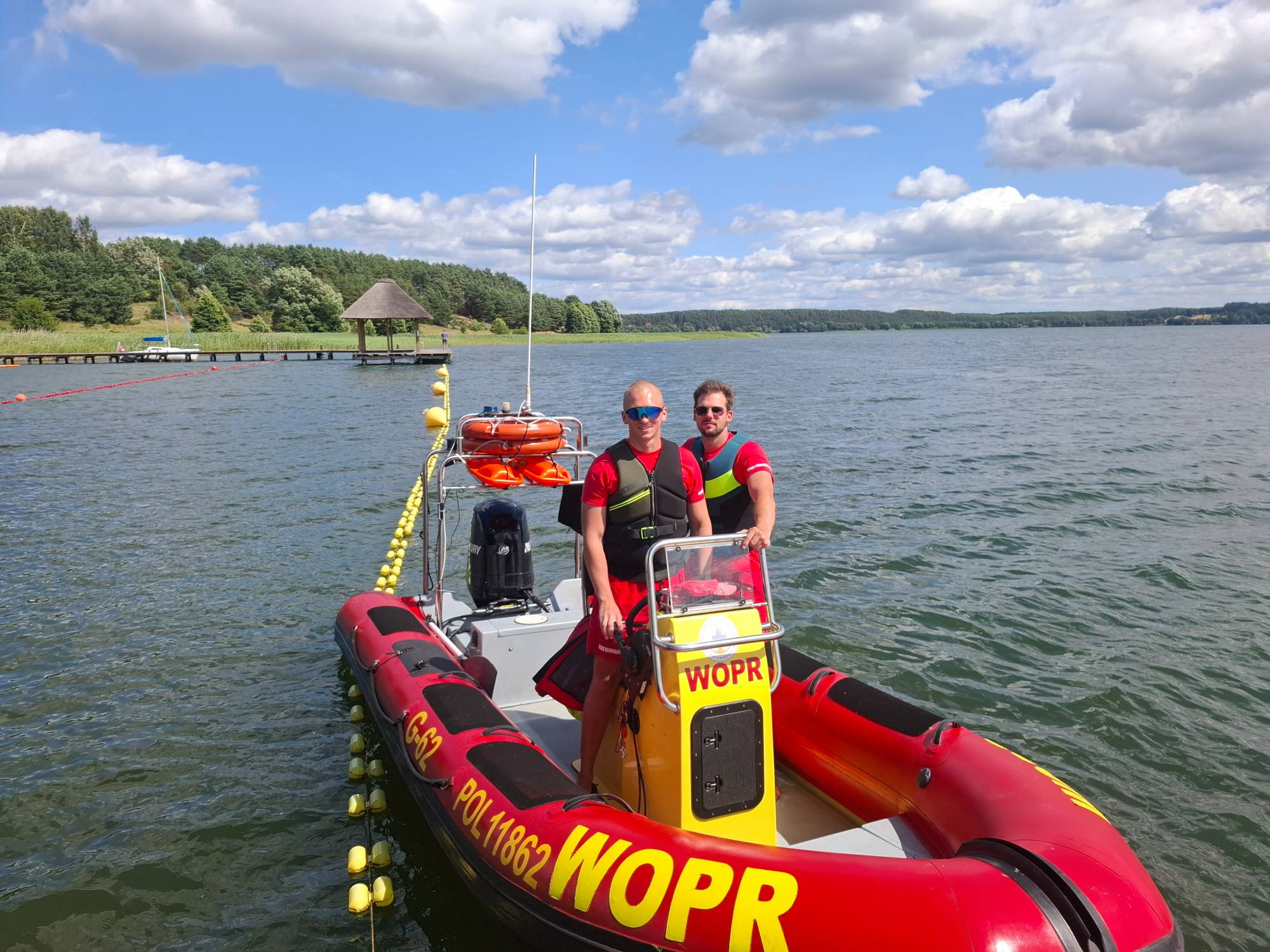 Dwaj ratownicy WOPR w czerwono-czarnych strojach i kamizelkach asekuracyjnych stoją na czerwono-żółtej łodzi motorowej na jeziorze. W tle widać pomost z drewnianą altaną krytą strzechą, żółte boje wyznaczające kąpielisko oraz błękitne niebo z chmurami