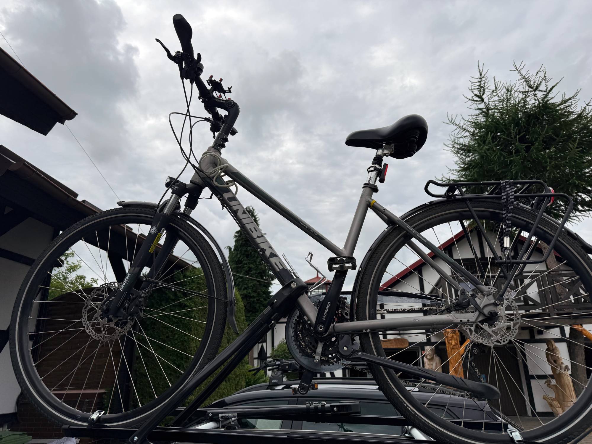 Szary rower trekkingowy marki Unibike zamontowany w uchwycie bagażnika na dachu czarnego samochodu. Zdjęcie zostało wykonane od dołu (z żabiej perspektywy) na tle zachmurzonego, szarego nieba oraz tradycyjnych domów z elementami muru pruskiego