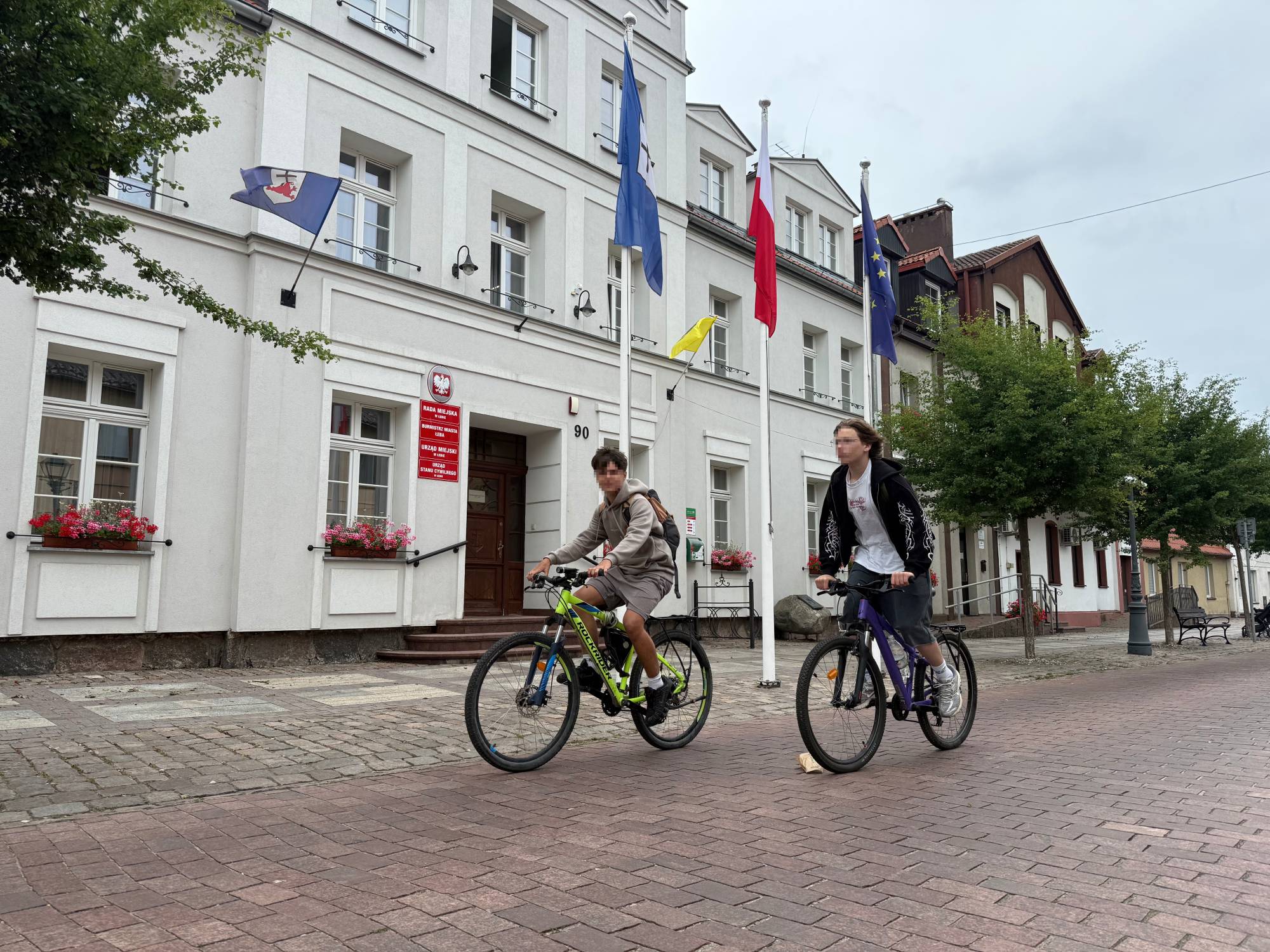 Dwóch nastolatków na rowerach górskich (jeden na jaskrawozielonym, drugi na fioletowym) jedzie po brukowanej ulicy przed białym, zabytkowym budynkiem Urzędu Miejskiego w Łebie. Przed wejściem do urzędu, na którym wiszą czerwone tablice informacyjne, stoją wysokie maszty z flagami Polski, Unii Europejskiej oraz miasta Łeby