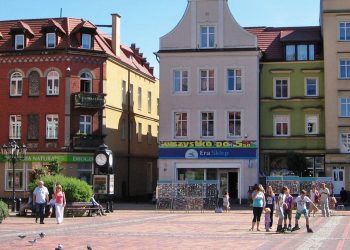 Zdjęcie przedstawia zabytkową pierzeję rynku w Chojnicach w słoneczny dzień. W tle widoczne są przylegające do siebie kolorowe kamienice: od lewej czerwona z cegły, jasnoszara z szyldem sklepu, zielona, żółta oraz błękitna. Na brukowanym placu przed budynkami spacerują ludzie, w tym grupa dzieci na rolkach, a po lewej stronie widoczne są ławki, miejski zegar i spacerujące gołębie