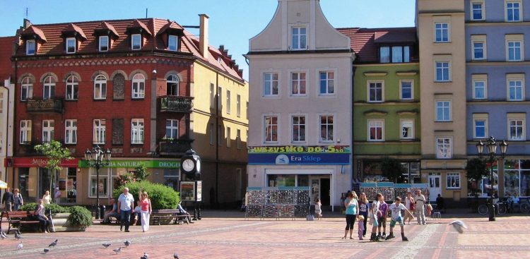 Zdjęcie przedstawia zabytkową pierzeję rynku w Chojnicach w słoneczny dzień. W tle widoczne są przylegające do siebie kolorowe kamienice: od lewej czerwona z cegły, jasnoszara z szyldem sklepu, zielona, żółta oraz błękitna. Na brukowanym placu przed budynkami spacerują ludzie, w tym grupa dzieci na rolkach, a po lewej stronie widoczne są ławki, miejski zegar i spacerujące gołębie