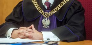 Zbliżenie na polskiego sędziego siedzącego przy stole sędziowskim. Osoba ta ma na sobie czarną togę z fioletowym żabotem i fioletowymi wykończeniami, co wskazuje na wydział karny. Na szyi sędziego spoczywa masywny, ozdobny złoty łańcuch z godłem Polski (orłem w koronie) i symbolem „RP”. Sędzia ma splecone dłonie oparte na dokumentach leżących na stole, a w tle widoczne jest oparcie czerwonego fotela