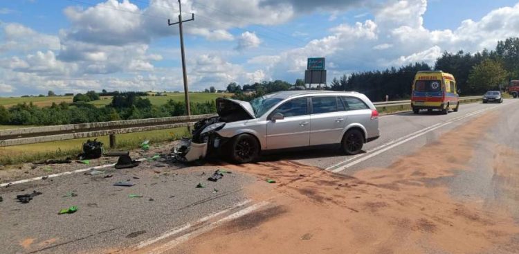 Srebrny samochód osobowy typu kombi z mocno rozbitym przodem i podniesioną maską stoi na zablokowanej drodze. Na jezdni wokół auta leżą rozrzucone odłamki i rozsypany sorbent, a w tle widać stojącą na poboczu żółtą karetkę pogotowia. Miejsce wypadku na drodze między Żukowem a Glinczem, w którym śmierć poniósł motocyklista