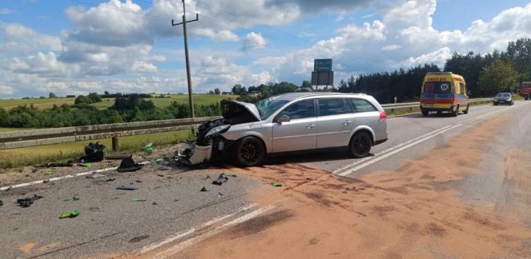 Srebrny samochód osobowy typu kombi z mocno rozbitym przodem i podniesioną maską stoi na zablokowanej drodze. Na jezdni wokół auta leżą rozrzucone odłamki i rozsypany sorbent, a w tle widać stojącą na poboczu żółtą karetkę pogotowia. Miejsce wypadku na drodze między Żukowem a Glinczem, w którym śmierć poniósł motocyklista