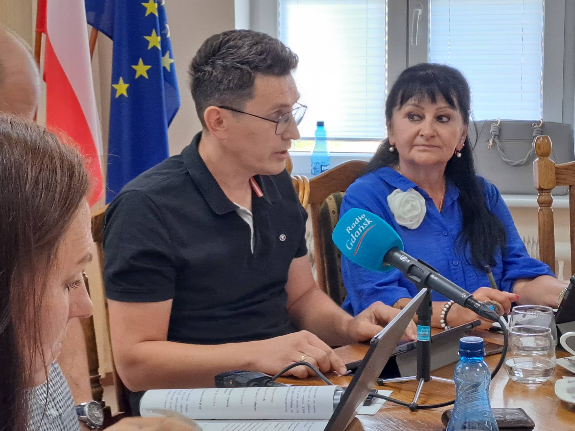 Mężczyzna w okularach i czarnej koszulce Polo przemawia przy stole konferencyjnym, na którym stoi laptop, dokumenty i mikrofon Radio Gdańsk. Obok niego siedzi kobieta w niebieskiej bluzce z ozdobnym białym kwiatem, a w tle wiszą flagi Polski i Unii Europejskiej. To rolnicy i samorządowcy z gminy Debrzno, który dyskutują na temat przyjętego planu ogólnego gminy, spierając się o miejsca wykluczone z zabudowy - w tym stawiania wiatraków