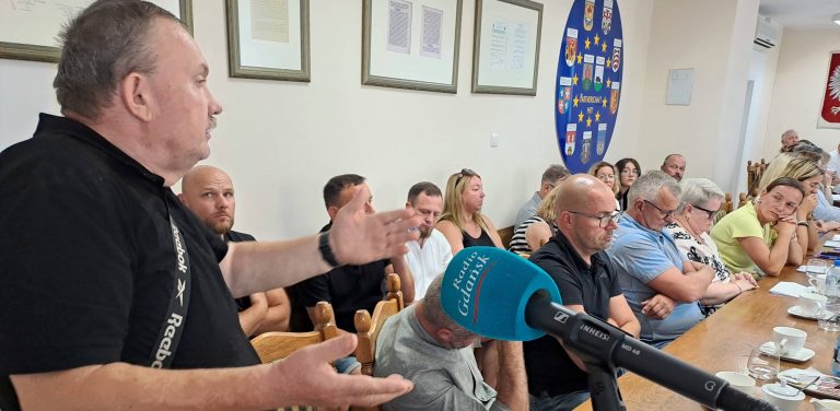 Mężczyzna w czarnej koszulce z logo Reebok przemawia z otwartymi dłońmi do mikrofonu Radio Gdańsk. W tle przy długim stole siedzą zgromadzeni uczestnicy spotkania. To rolnicy oraz samorządowcy z gminy Debrzno, gdzie doszło do konfliktu związanego z przyjętym planem ogólnym dla gminy. Rolnicy chcą powiększenia stref przeznaczonych pod lokalizacje wiatraków