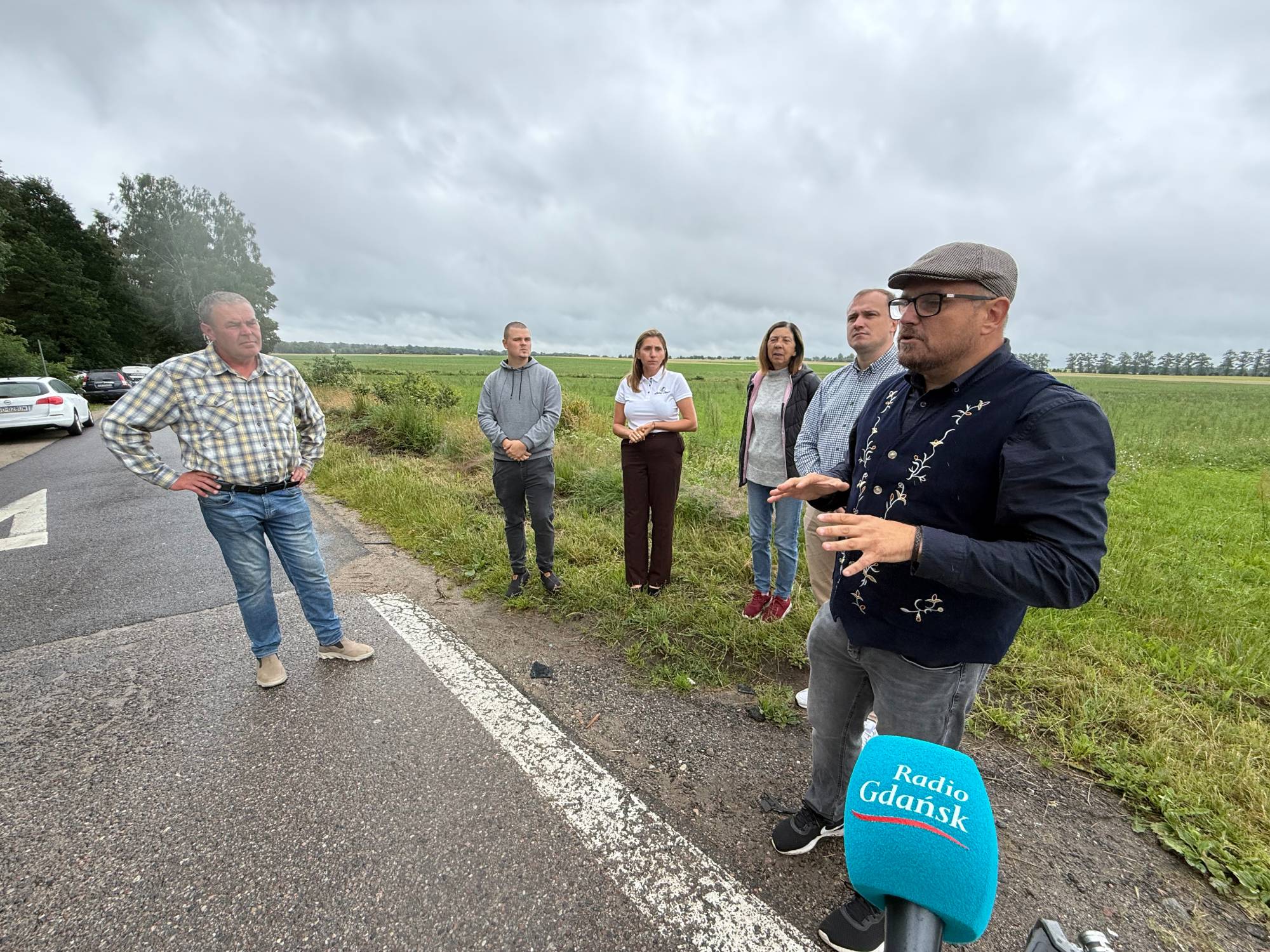 Mężczyzna w okularach, kaszkiecie i ciemnej kamizelce z jasnym haftem mówi i gestykuluje, trzymając dłonie przed sobą. W prawym dolnym rogu widoczny jest turkusowy mikrofon z logo „Radio Gdańsk”. W tle na poboczu drogi stoi grupa pięciu osób słuchających wypowiedzi, a za nimi rozciąga się zielone pole pod zachmurzonym niebem. Przedsiębiorcy tłumaczą reporterowi, że ich biznesy ucierpiały z powodu błędu GPS, który wykluczył z map nawigacyjnych starą drogę DK6
